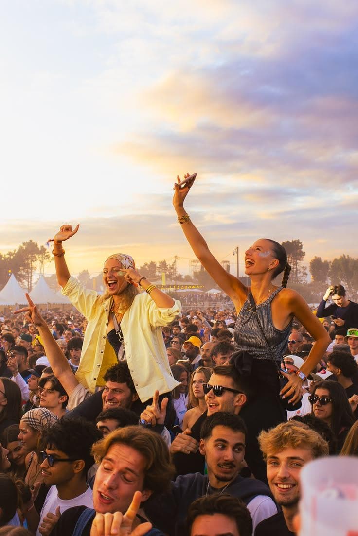 Two friends up on their friends' shoulders, arms raised, in a festival crowd at sunset